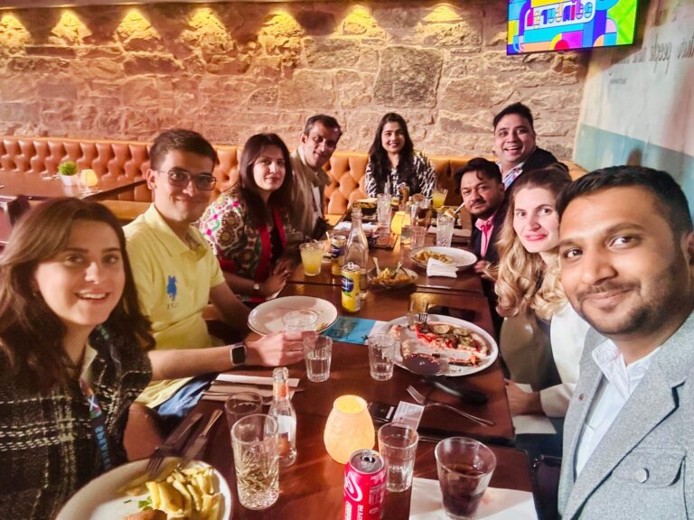 a group of people sit around a restaurant table enjoying a meal together, smiling for a group photo in a cozy setting with stone walls and warm lighting.