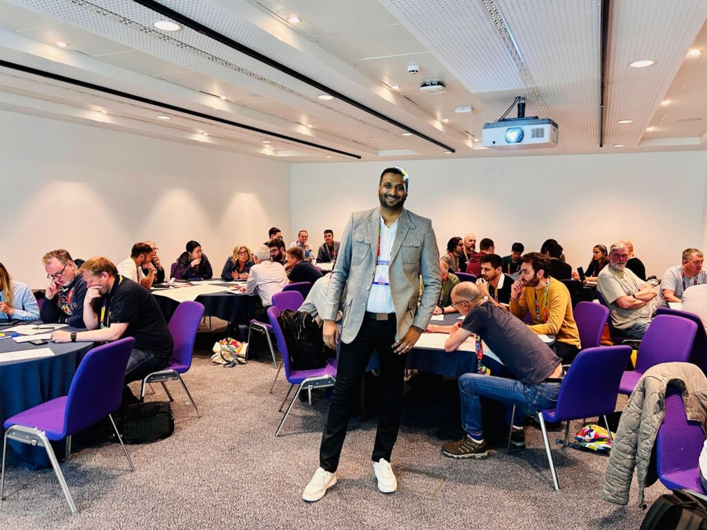 rahul parwal posing for a photo in the centre of a conference room where several groups of people are engaged in collaborative discussions and activities at round tables.