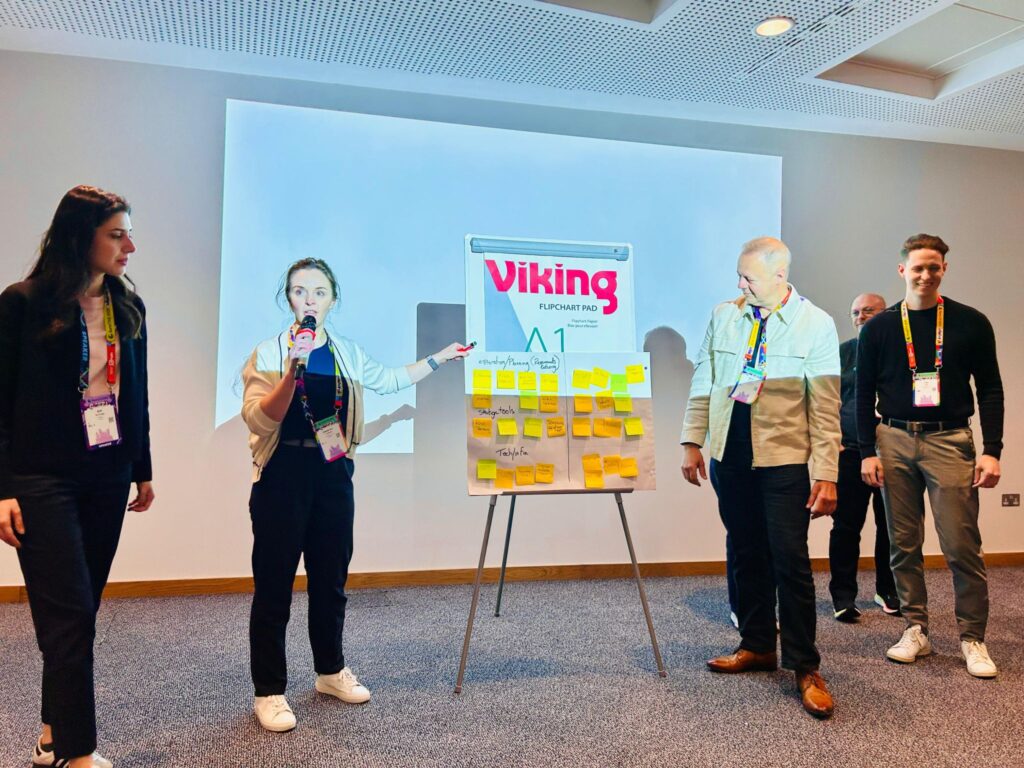a group presenting their work during a conference session. one person is speaking and pointing to a flip chart covered with sticky notes, while the others stand nearby listening.