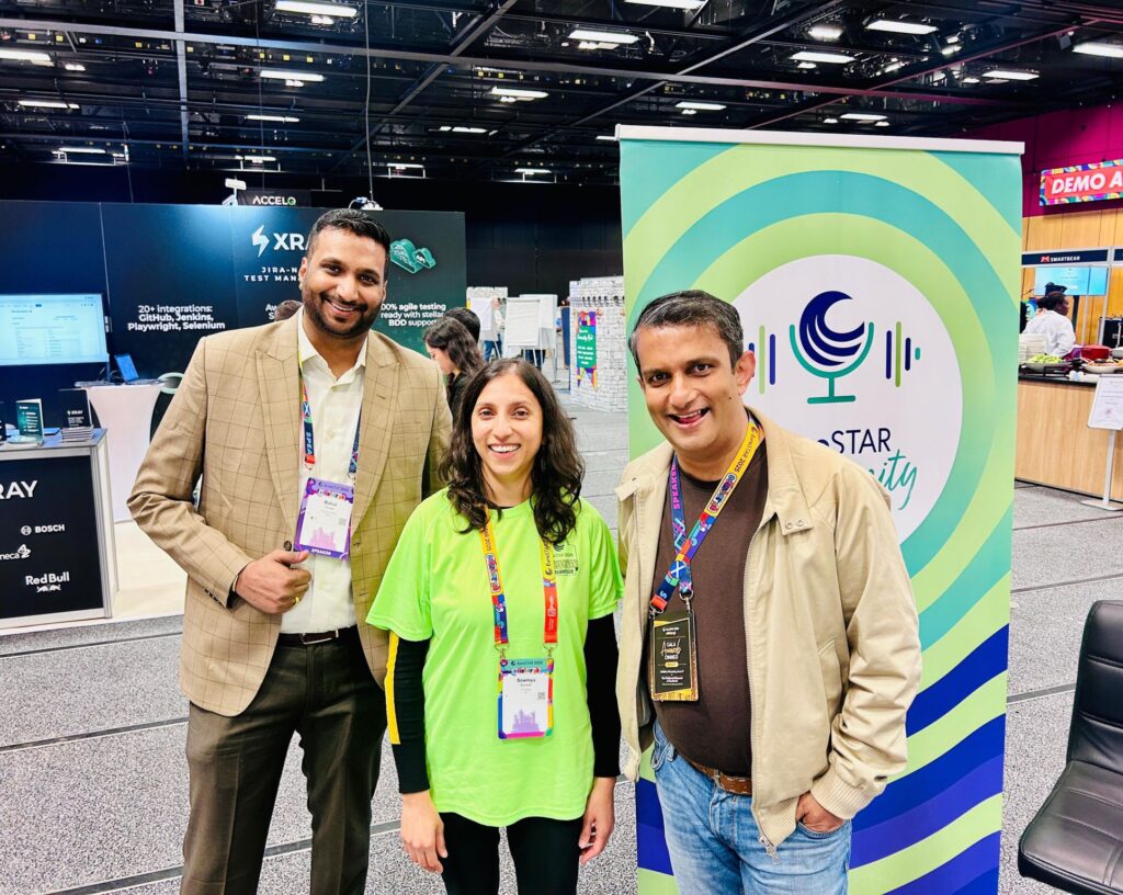 three conference attendees smiling together in front of a podcast community banner and exhibition booths.