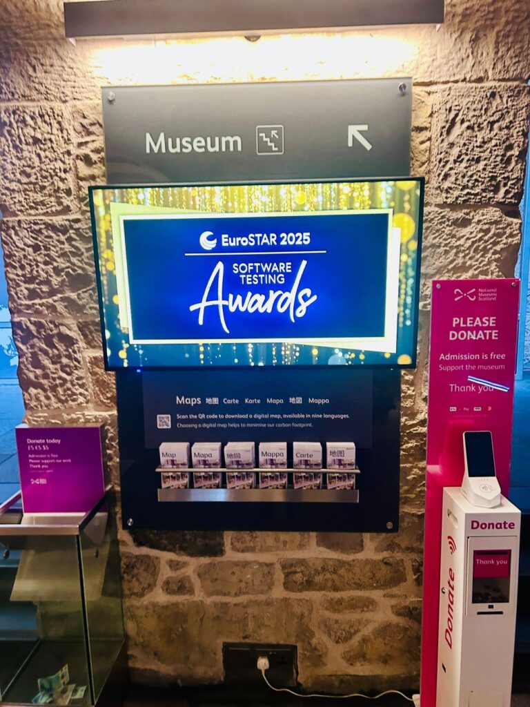 a display inside a museum showing the EuroSTAR 2025 software testing awards on a screen, with donation stands and informational materials nearby.
