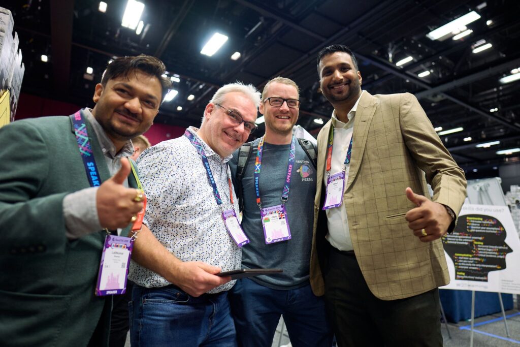 a group of four smiling individuals posing together at a conference event, wearing name badges and lanyards, with two giving thumbs up gestures, in a brightly lit exhibition hall with high ceilings and display booths in the background.