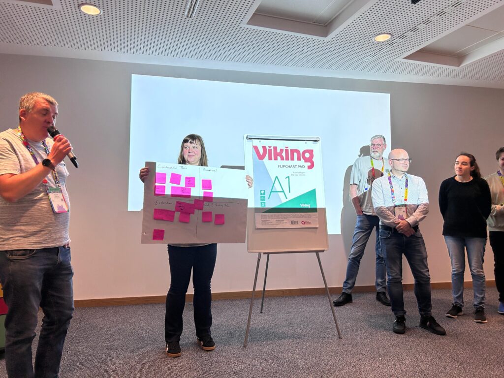 a group of people presenting during a workshop, with one person speaking while another holds a board covered with pink sticky notes, standing beside a flip chart.