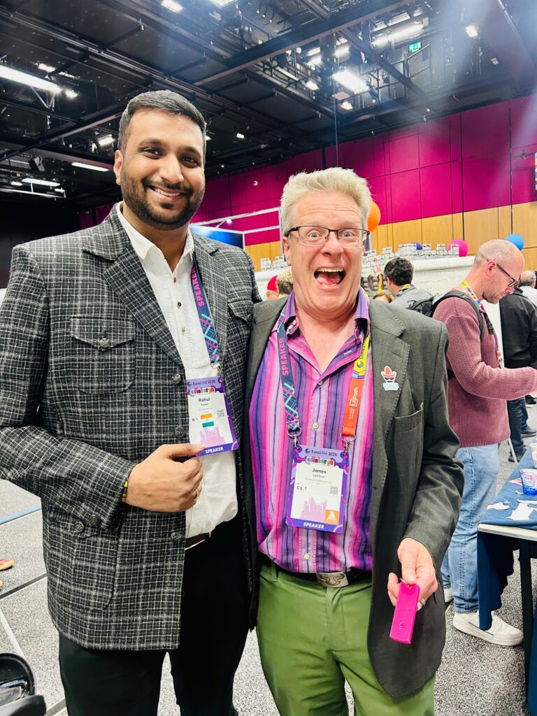 Two people smiling and posing for a cheerful photo at a conference event, with conference booths and attendees visible in the background.