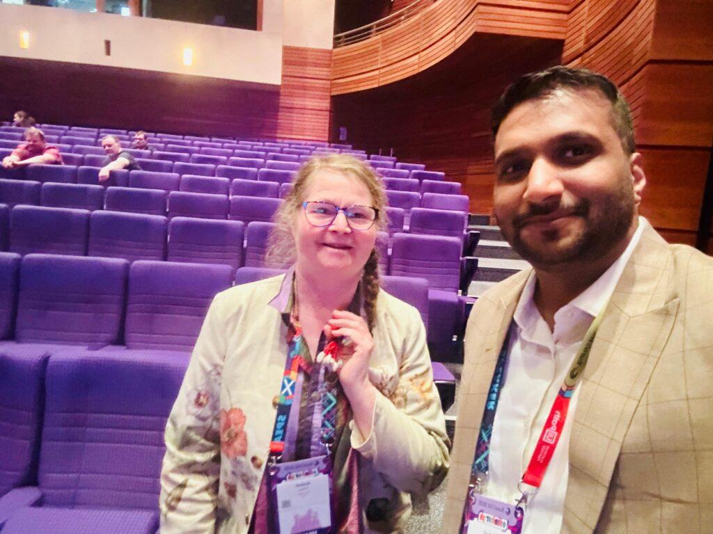 Two people smiling and posing for a photo inside an auditorium with rows of purple seats and a few people seated in the background.