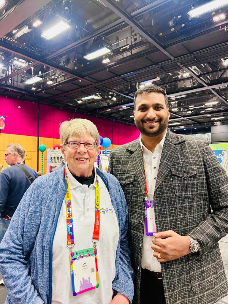Two people smiling and posing together at a conference venue, with colorful booths and other attendees in the background.